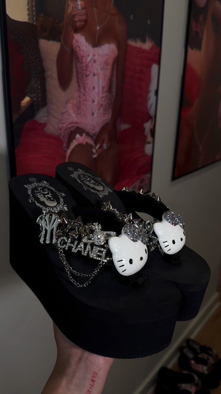 A hand holding black platform flip flops decorated with Hello Kitty charms and silver chains.