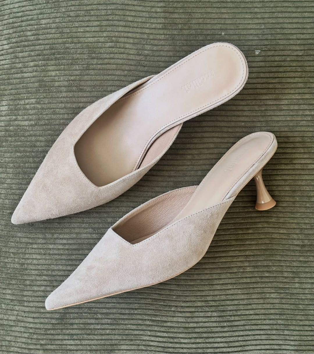 Nude suede pointed toe mule heels resting on a green corduroy textured background.