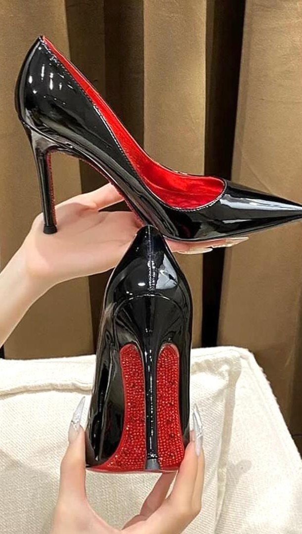 Hands holding black patent leather high heels with distinctive red soles and red interiors.
