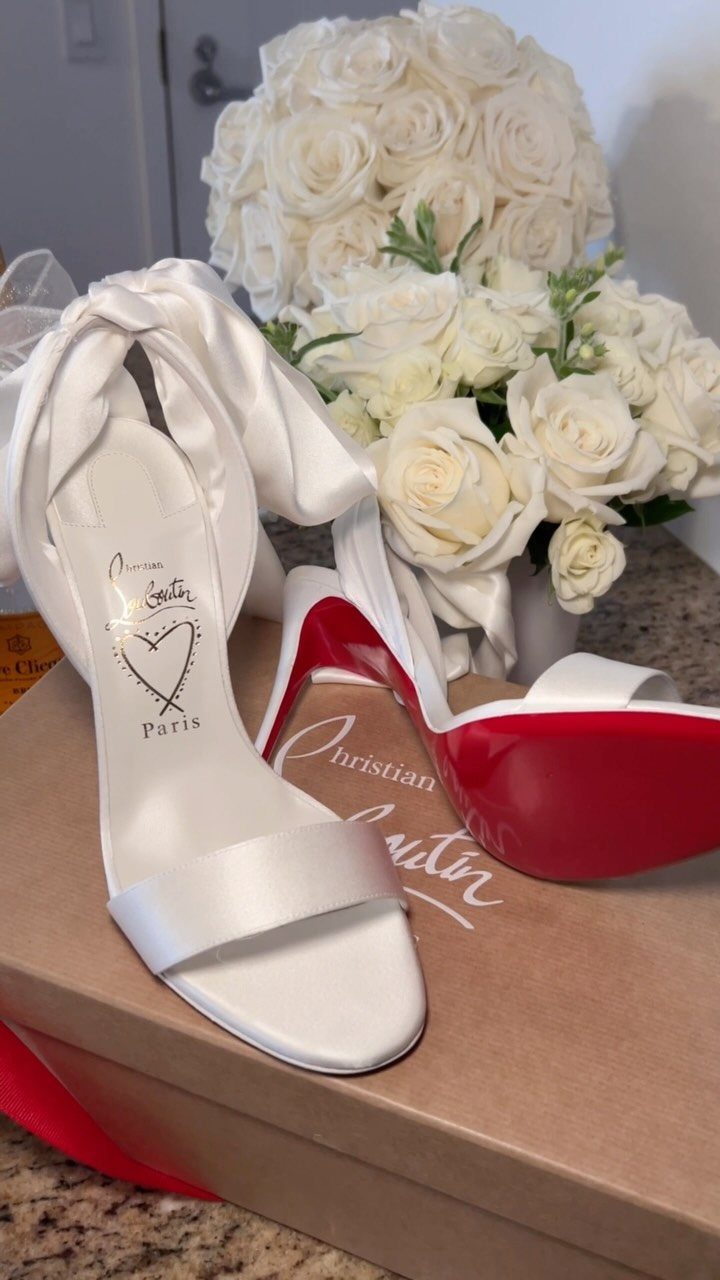 White satin high-heeled sandals with bright red soles resting on a brown box near white roses.