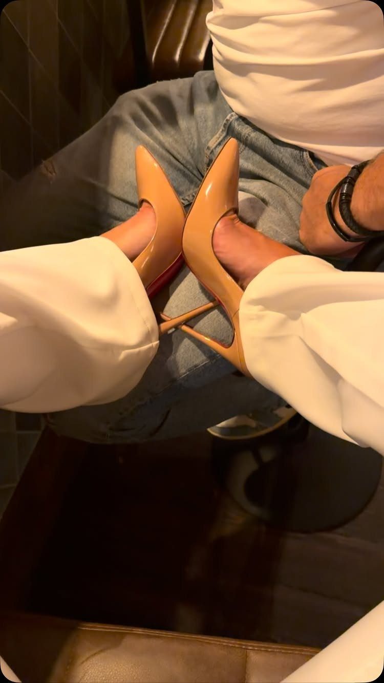 Beige patent leather high heels with red soles worn by a person in denim jeans.