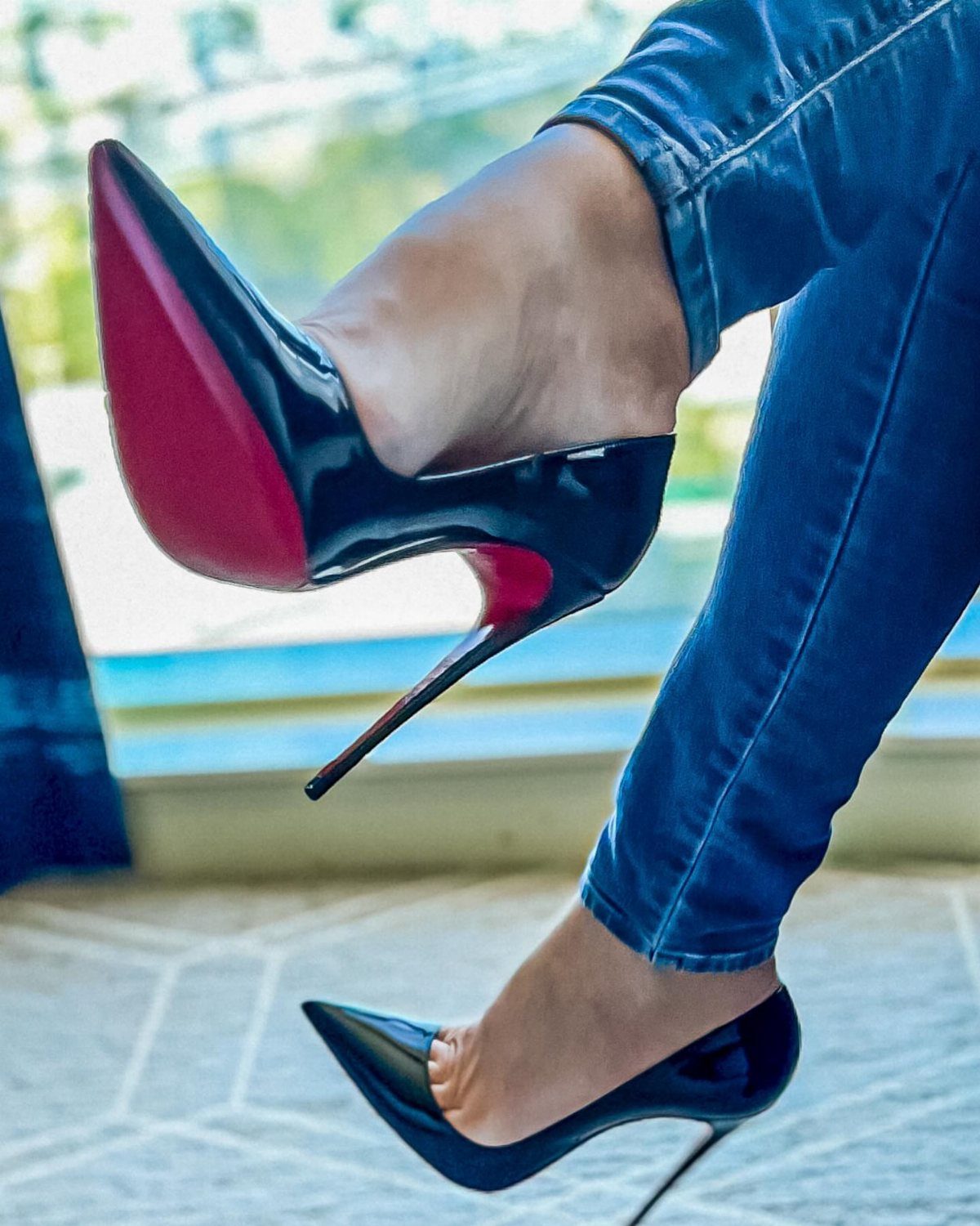 Black patent leather high heels with red soles worn by a person in blue jeans.