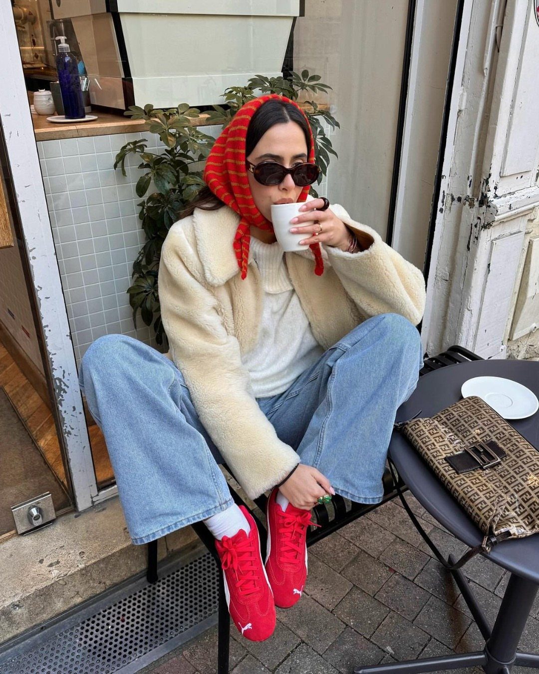Woman in cream jacket, blue jeans, and red sneakers sitting at an outdoor cafe table.
