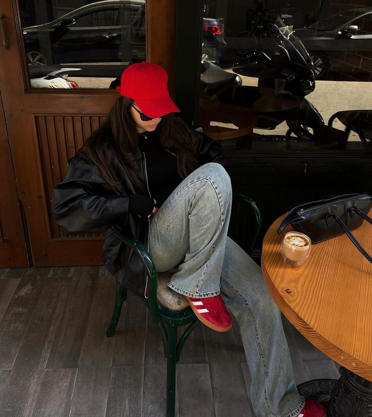Woman wearing a black leather jacket, red cap, blue jeans, and red sneakers sitting outdoors.