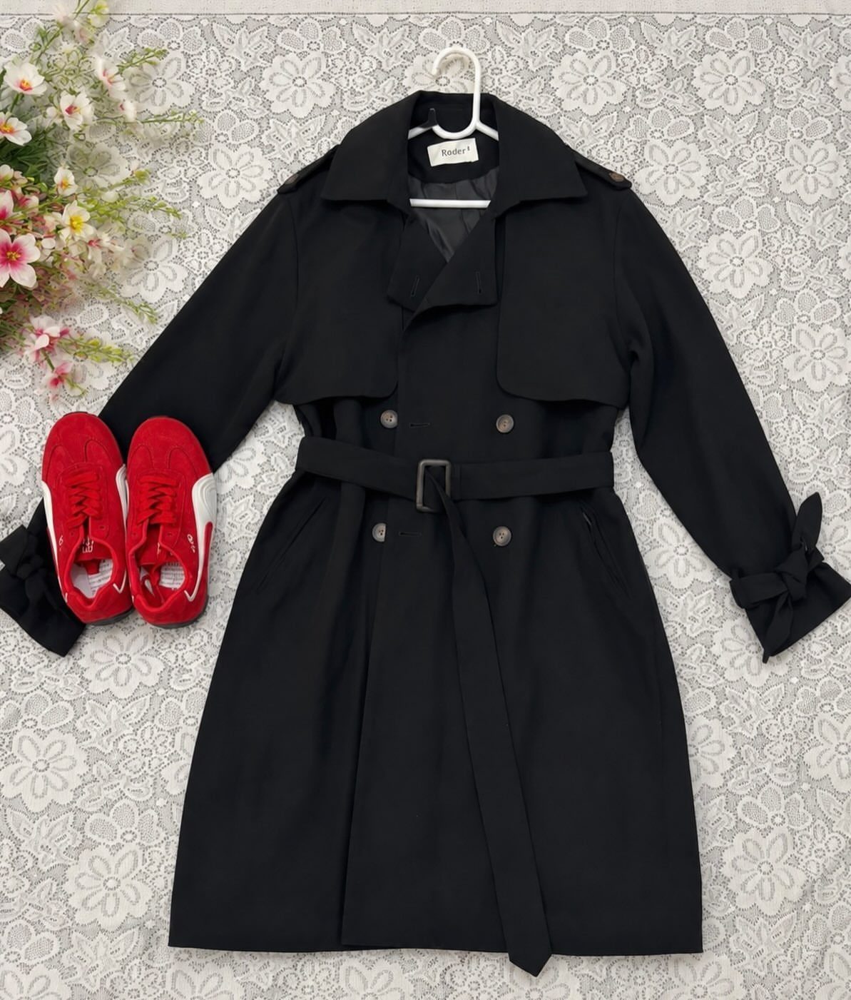 Black belted trench coat and red sneakers laid out on a white floral patterned background.