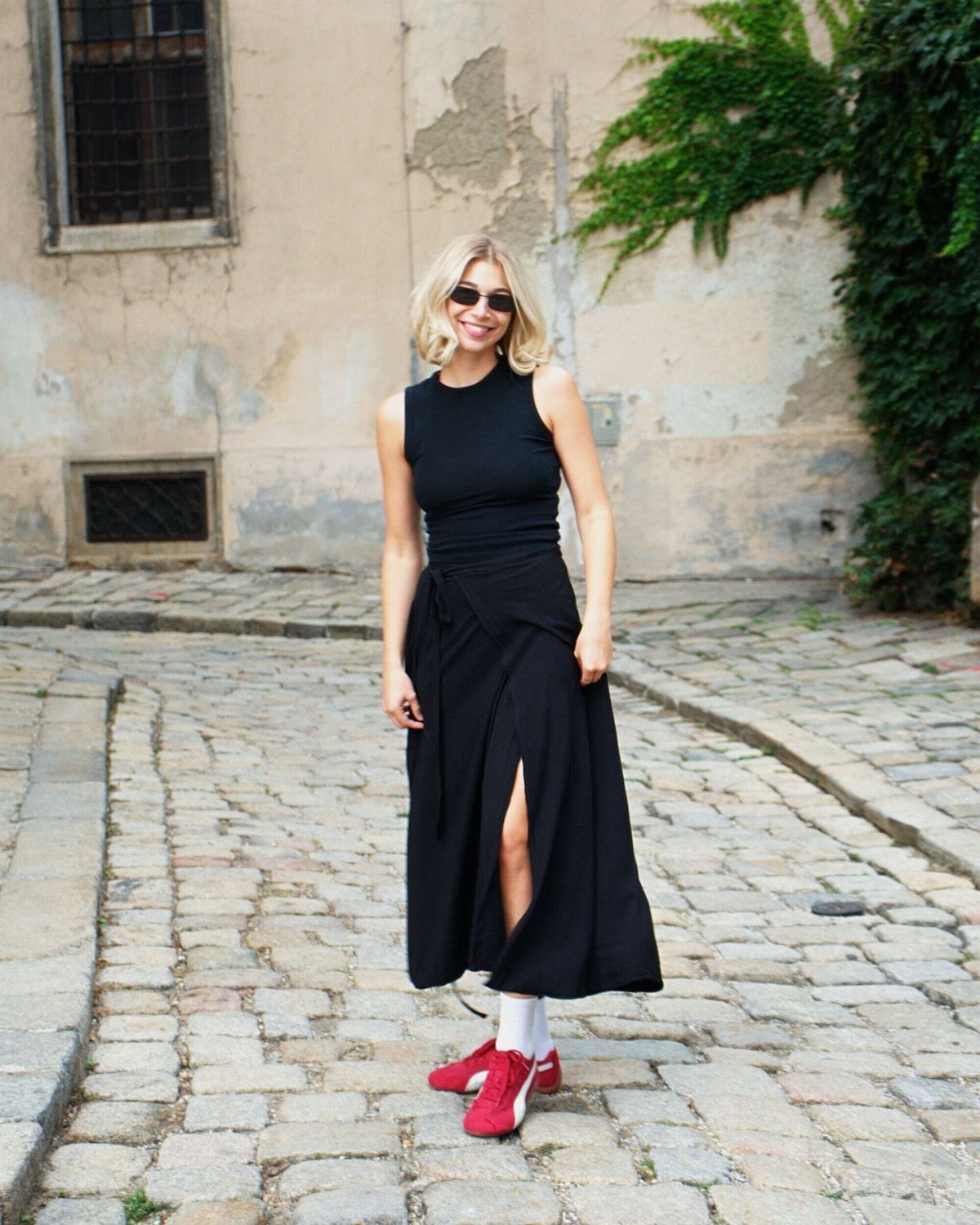 A blonde woman wearing a black midi skirt and red sneakers standing on a cobblestone street.