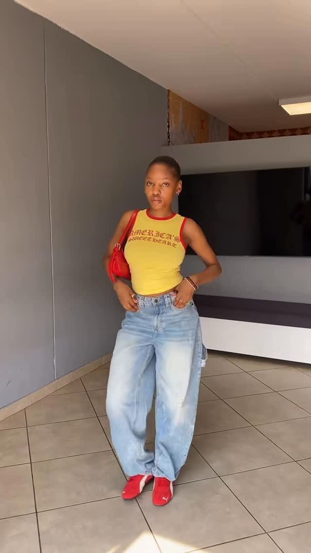 Woman in a yellow tank top, baggy blue jeans, and red sneakers standing indoors.