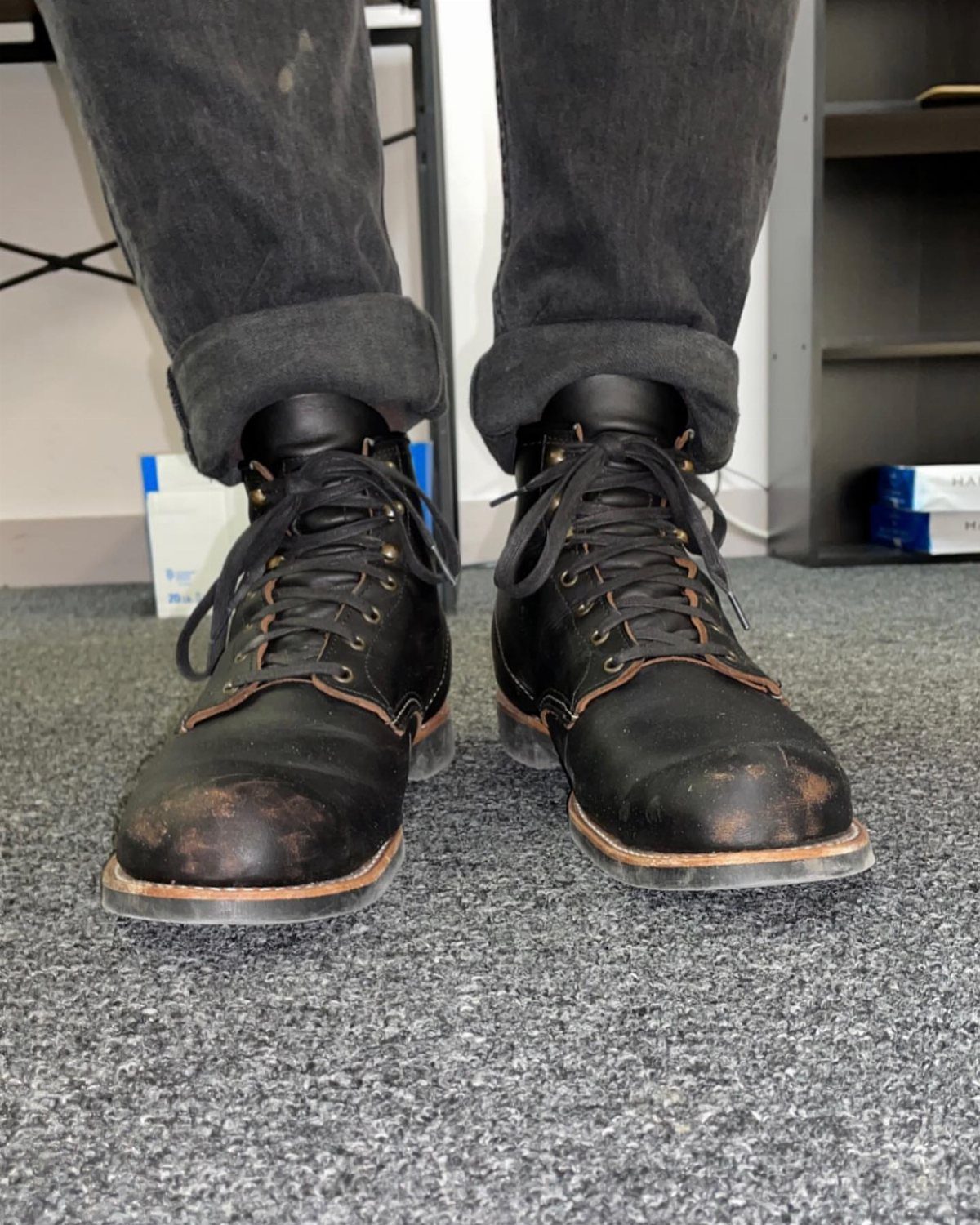 Black leather lace-up boots with scuffed toes worn with cuffed dark grey jeans.