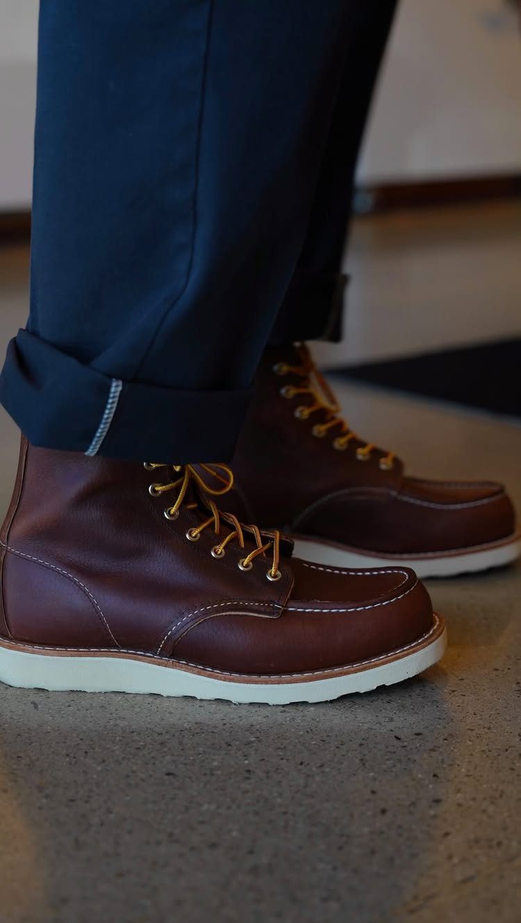 Brown leather moc-toe boots with white soles worn by a person in navy trousers.