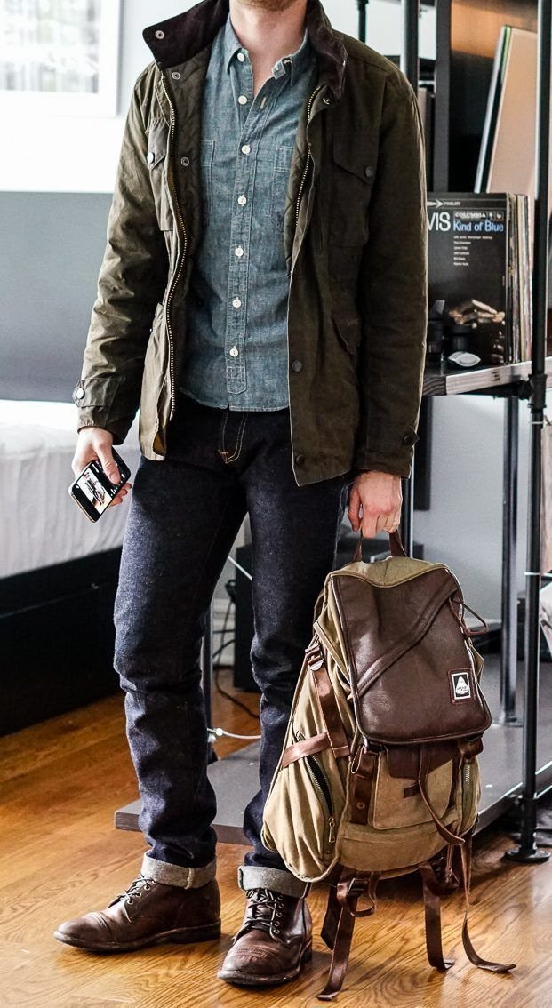 Man wearing an olive jacket, denim shirt, dark jeans, and brown leather boots standing by a backpack.