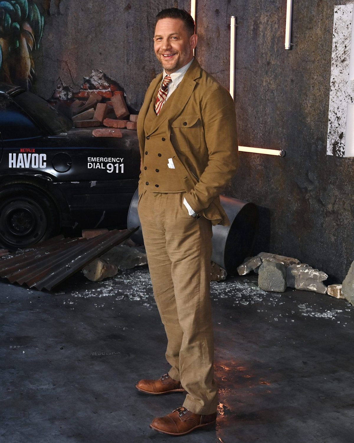 Man in a tan suit and brown leather boots smiling in an industrial setting.