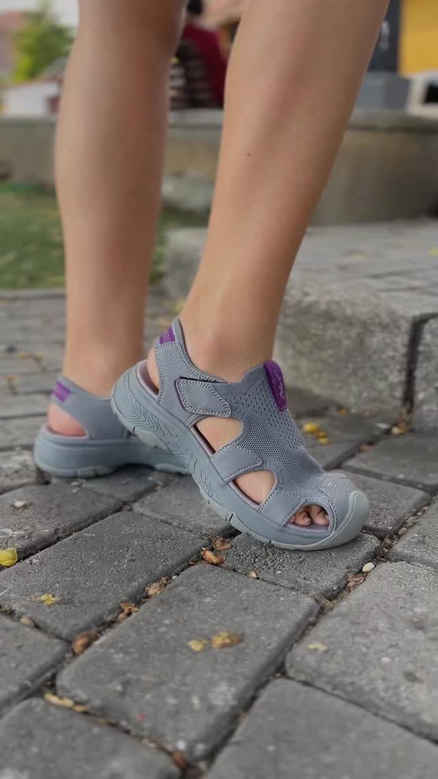 Grey closed-toe walking sandals with purple accents worn on a person's feet on a stone path.