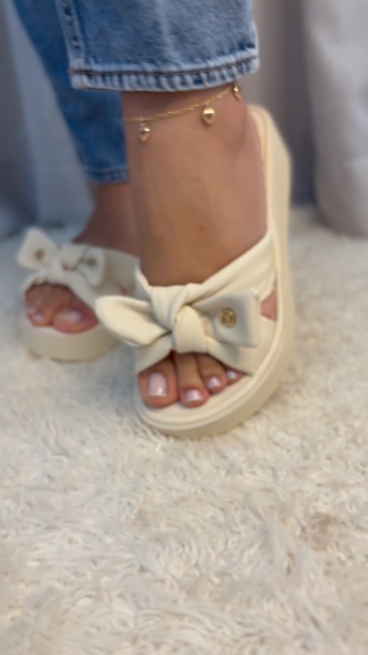 Cream platform sandals with large decorative bows worn by a person on a white rug.
