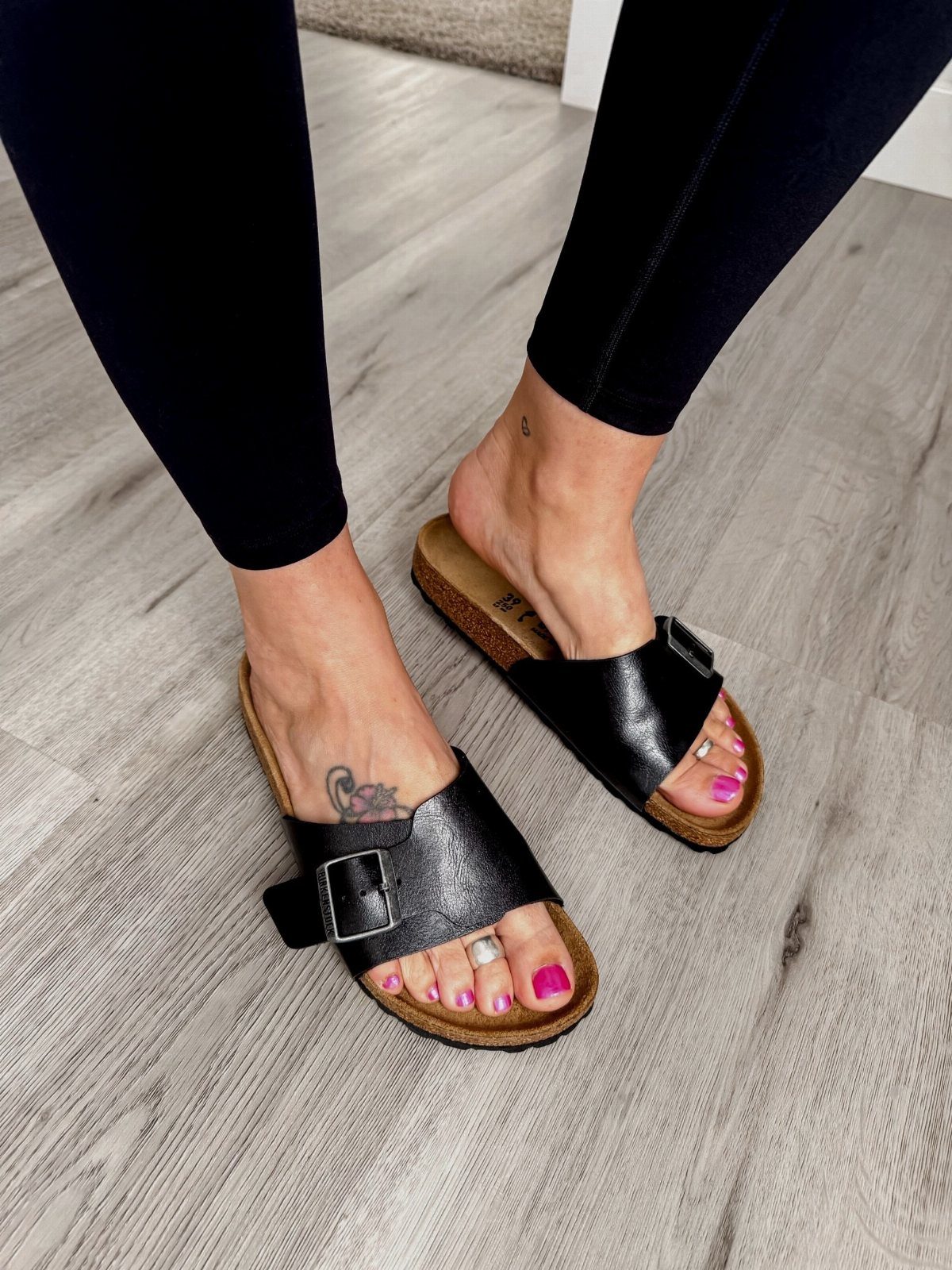 Person wearing black leather slide sandals with cork soles on a grey wood floor.
