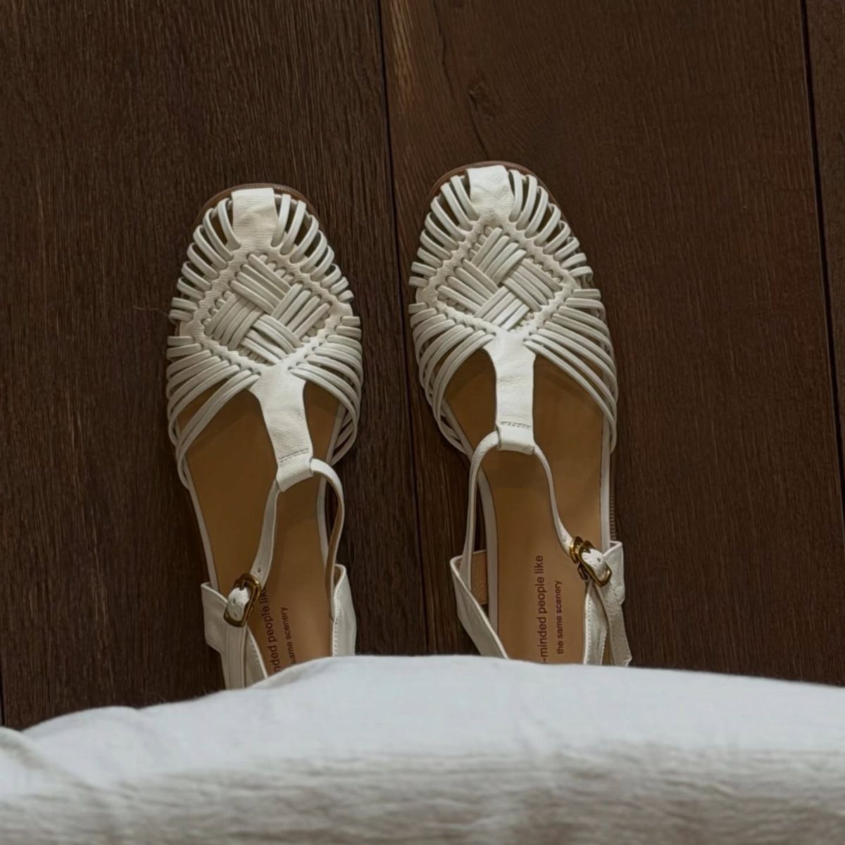 White woven leather sandals with ankle straps resting on a dark wooden floor surface.