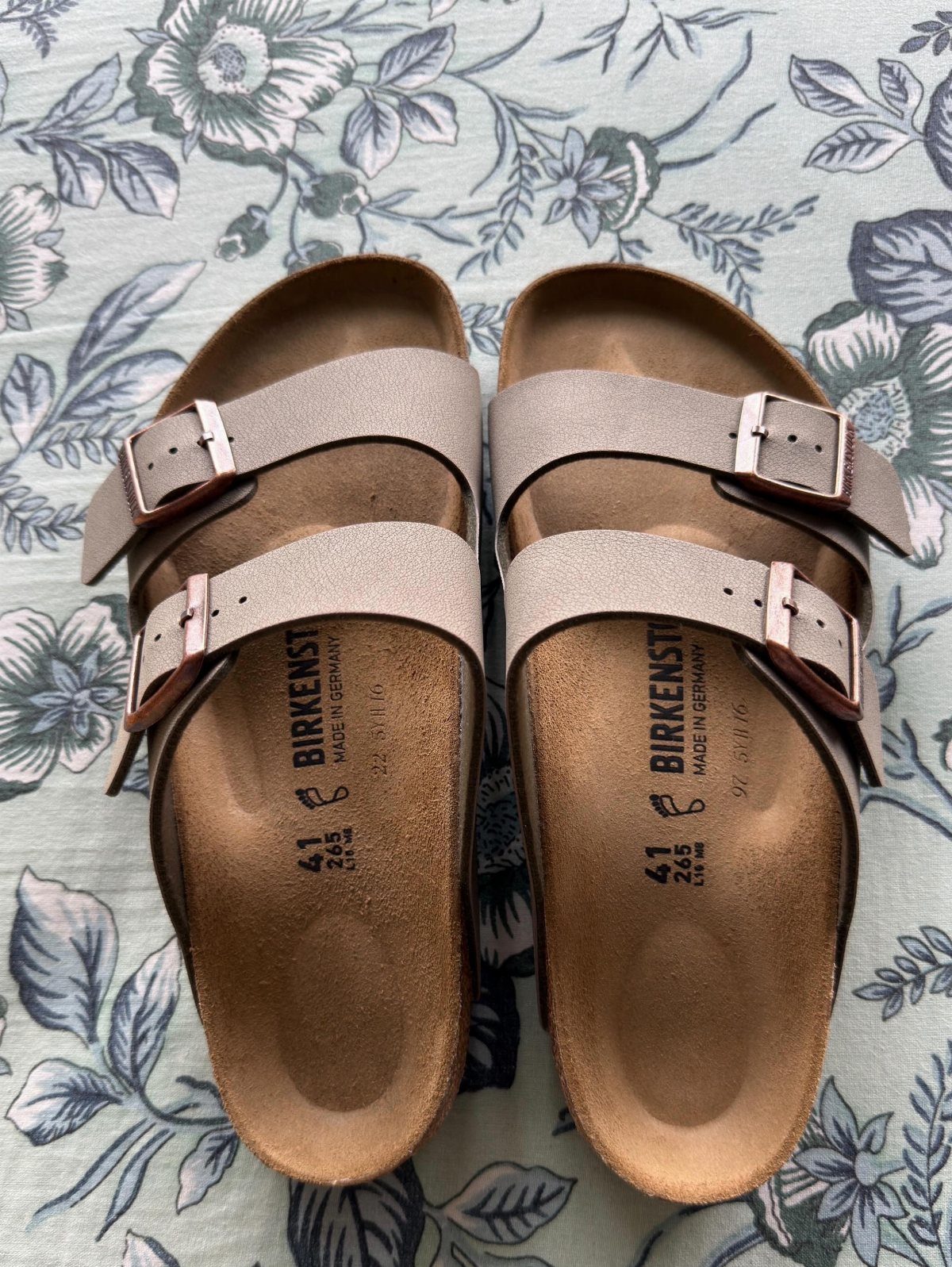Tan two-strap Birkenstock sandals resting on a blue and white floral patterned fabric background.