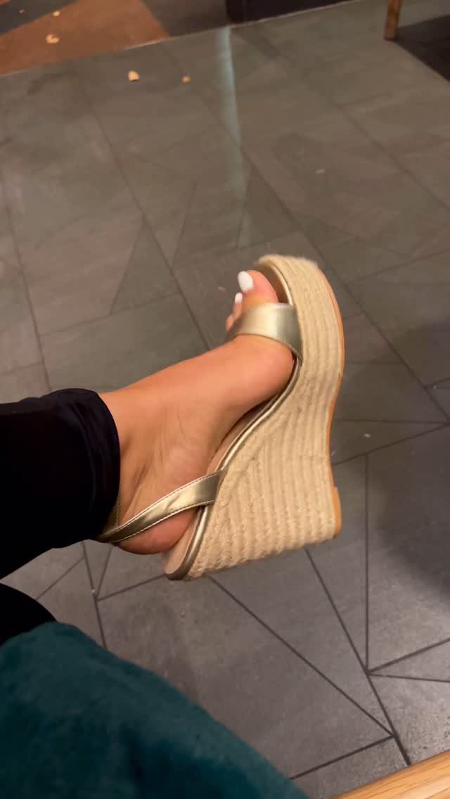 A foot wearing a gold metallic wedge sandal with a woven platform sole on a dark floor.