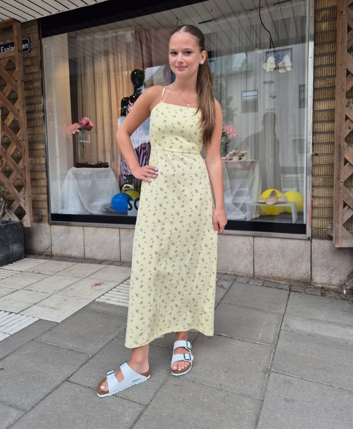Young woman in a yellow floral dress wearing white two-strap slide sandals standing outdoors.