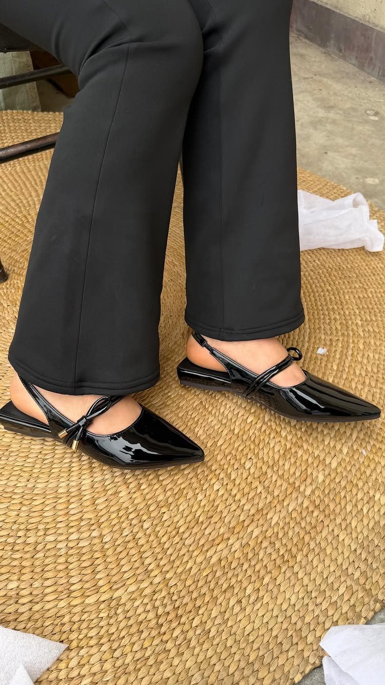 Black patent leather pointed-toe flats with thin ankle straps worn by a person in black trousers.