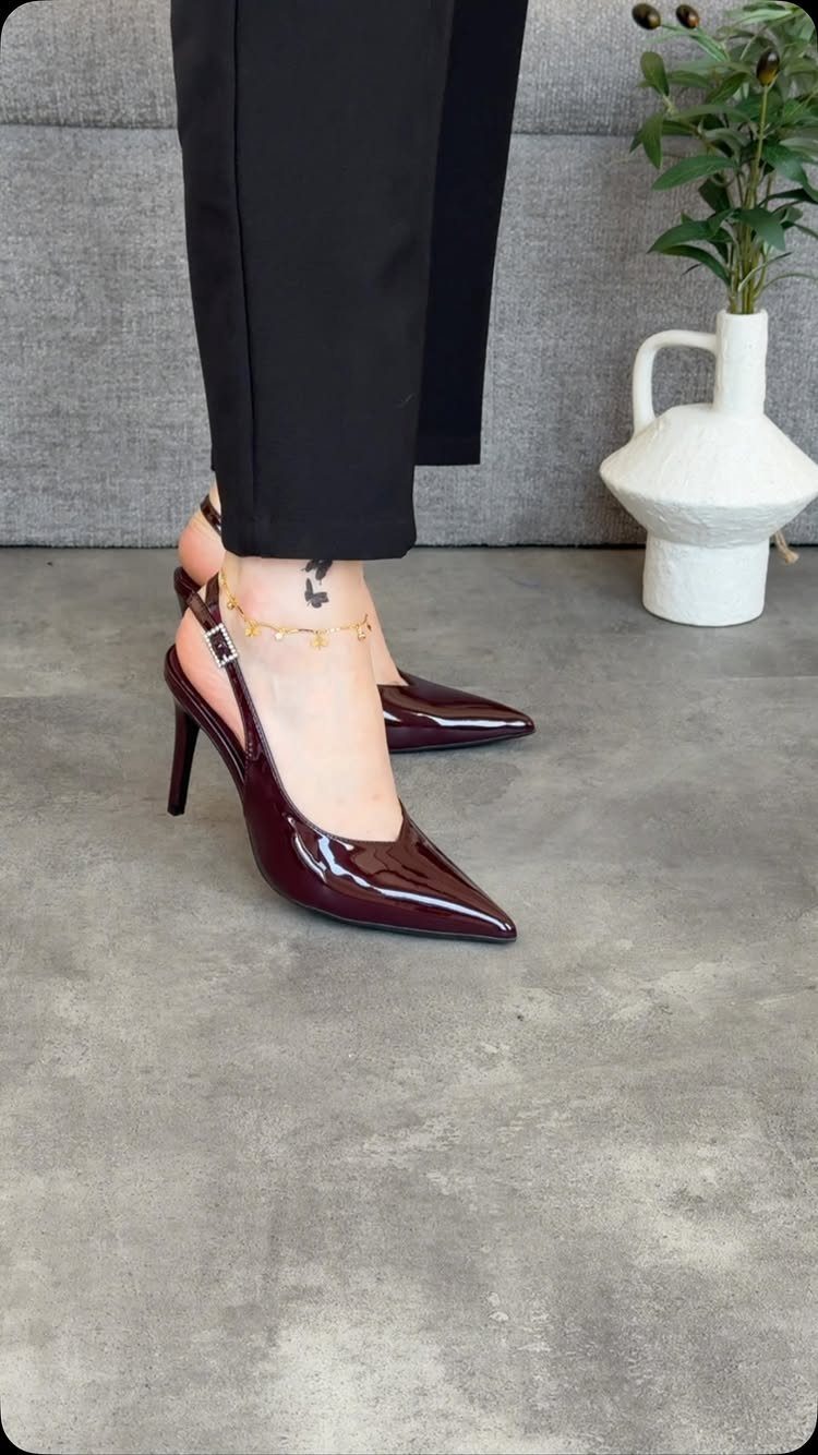 Burgundy patent leather pointed-toe stiletto pumps with ankle straps worn by a person in black trousers.