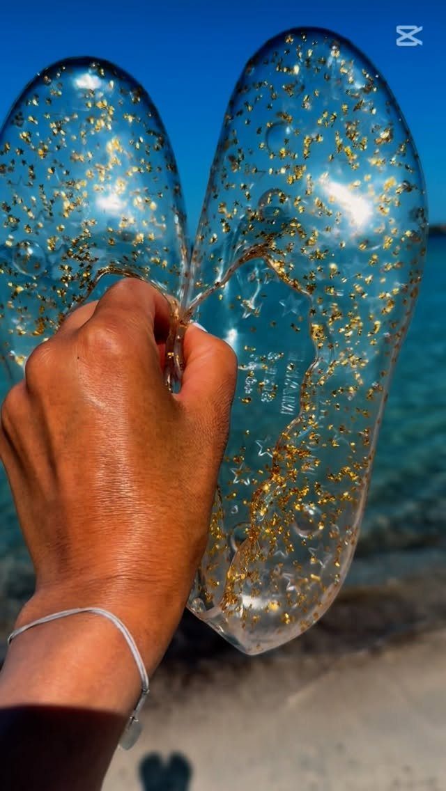 A hand holding up clear glittery sandals against a blue sky and ocean background.