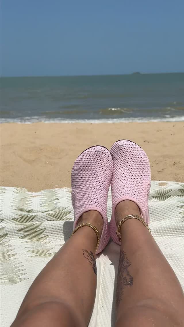 Pink crochet slip-on shoes worn by a person resting on a beach towel near the ocean.