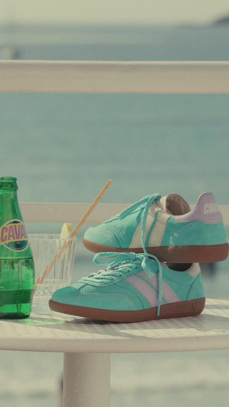 Teal and lavender sneakers resting on a white table near a green bottle by the ocean.
