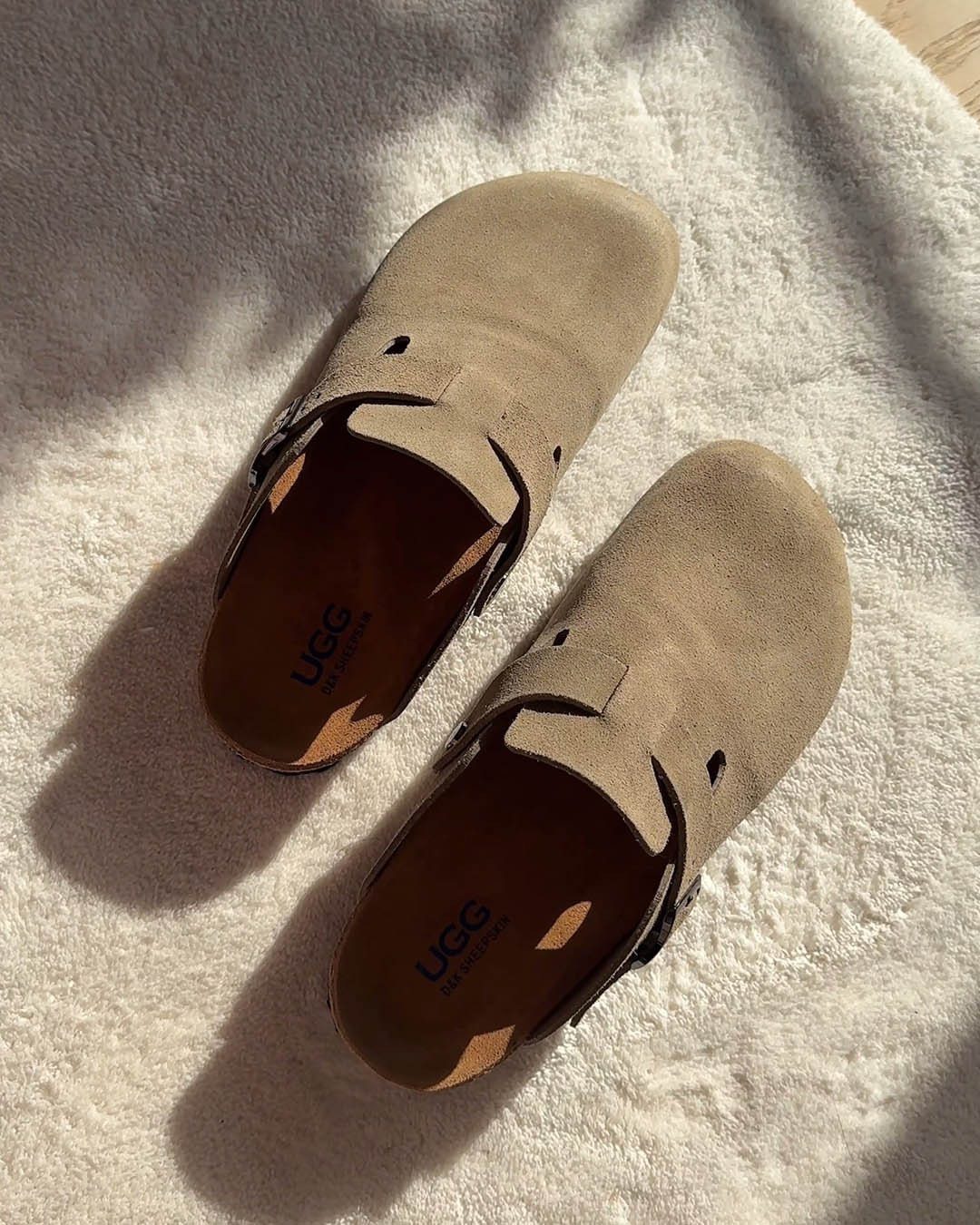 Tan suede UGG clogs with black straps resting on a white textured surface.