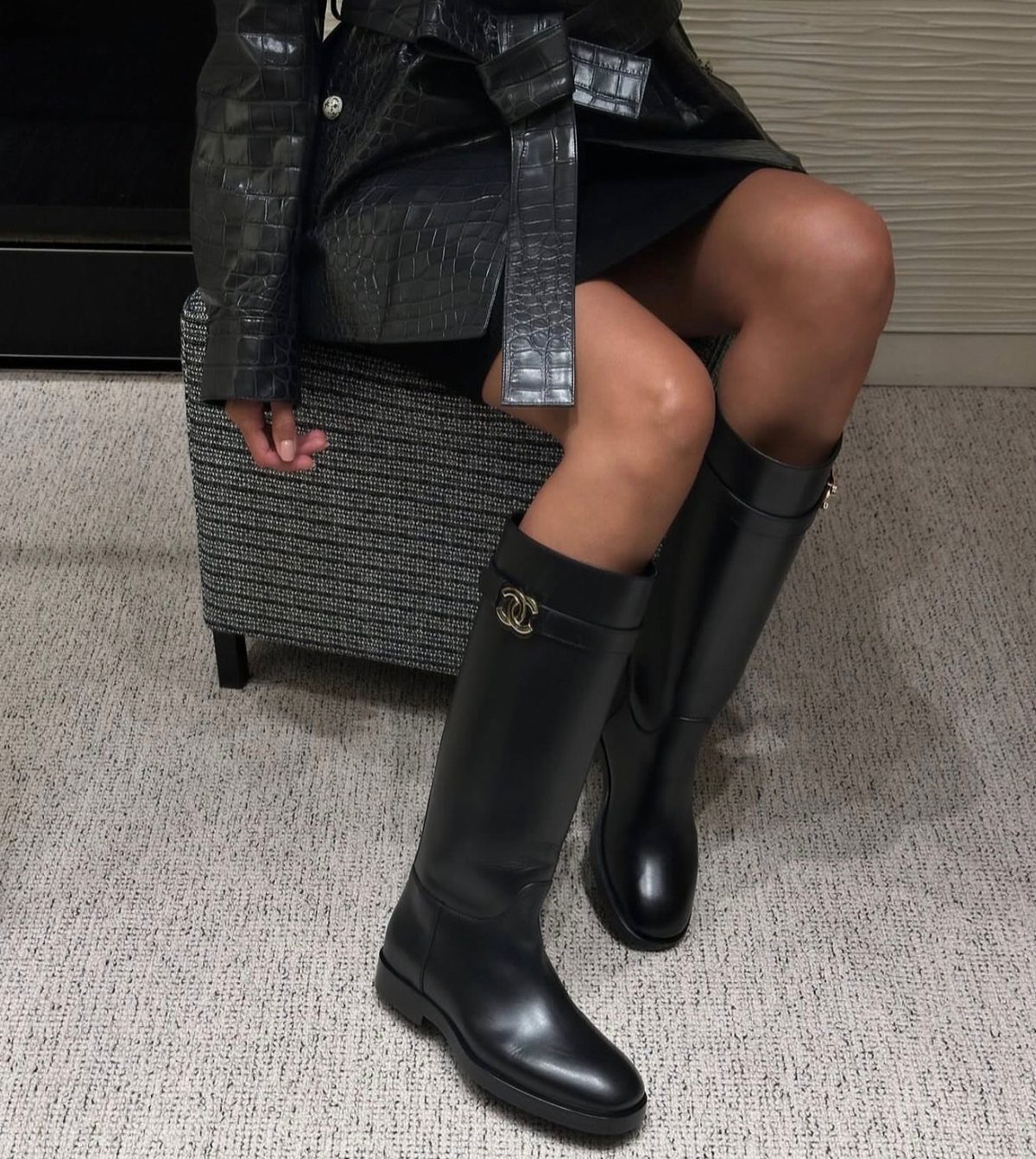 Black knee-high leather boots with gold hardware worn by a person sitting on an ottoman.
