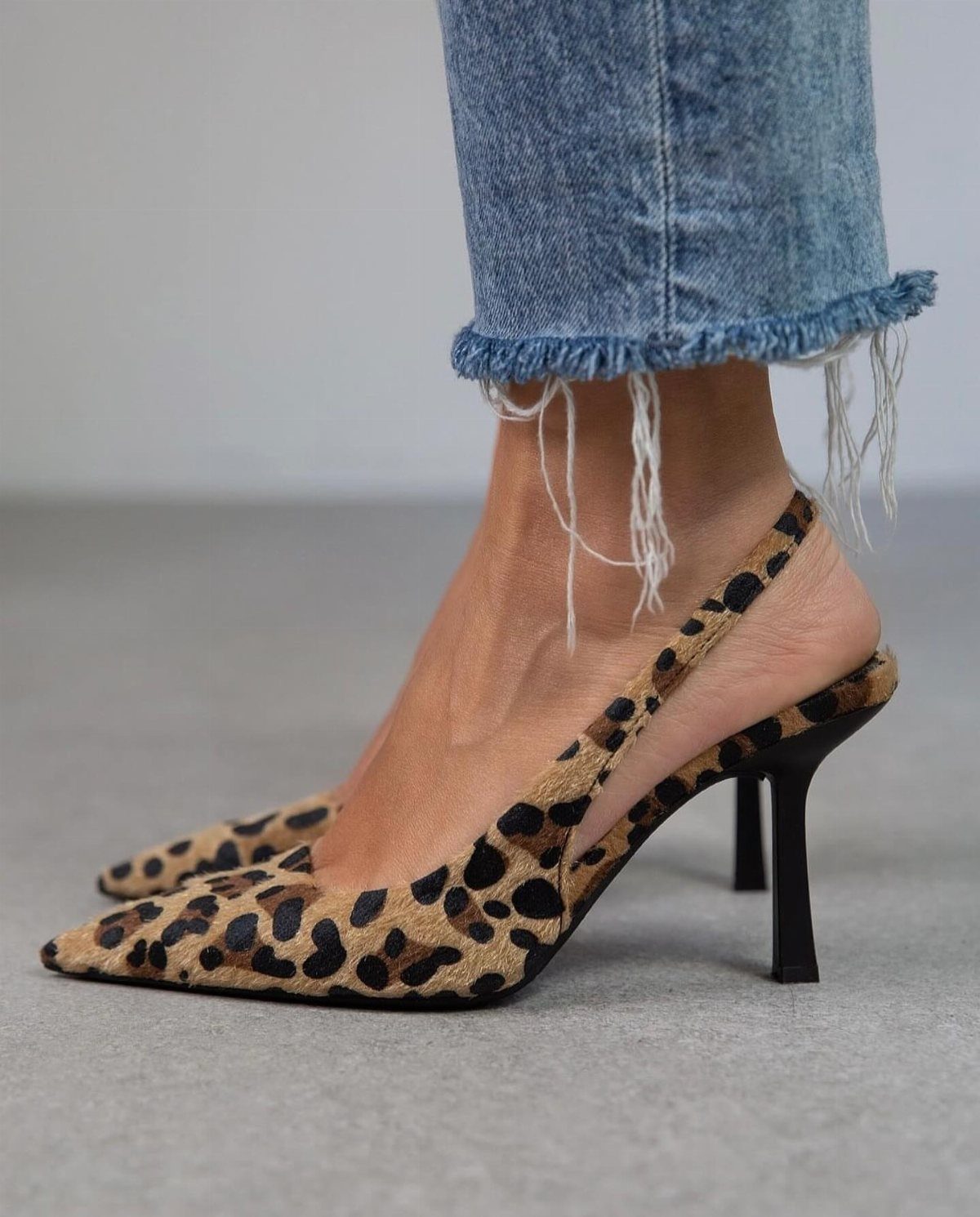 Leopard print pointed-toe slingback heels worn with frayed blue denim jeans on a neutral floor.