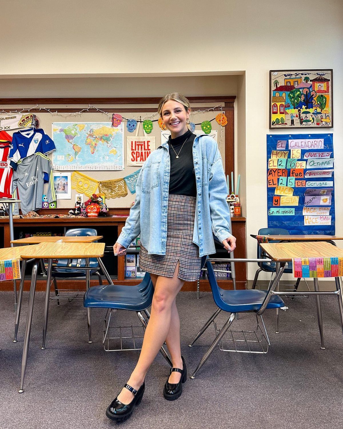 Smiling woman in a classroom wearing a denim jacket, patterned skirt, and black Mary Jane shoes.