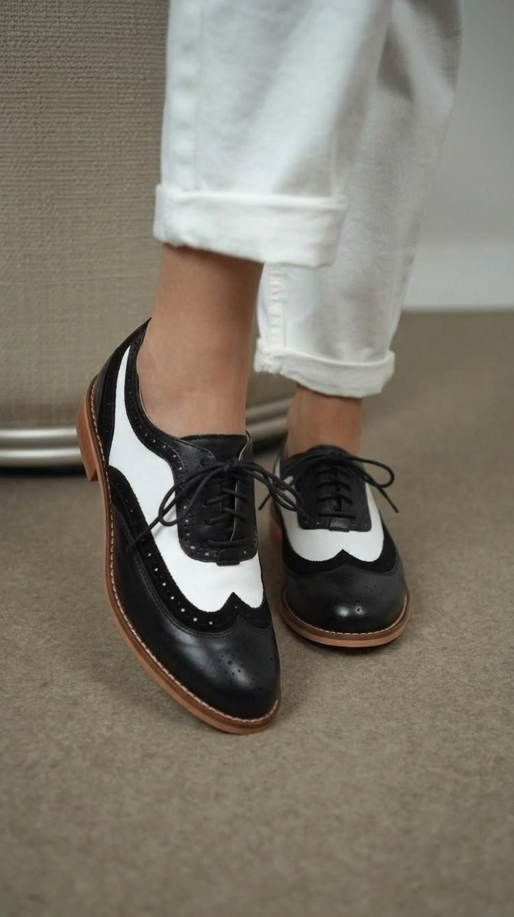 Black and white wingtip spectator shoes worn with white cuffed trousers on a neutral floor.