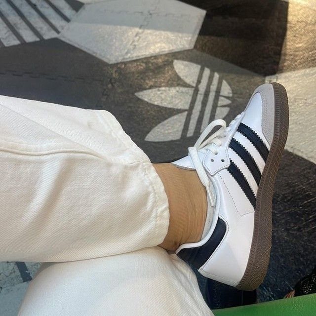 White Adidas Samba sneakers with black stripes worn with white trousers on a patterned floor.