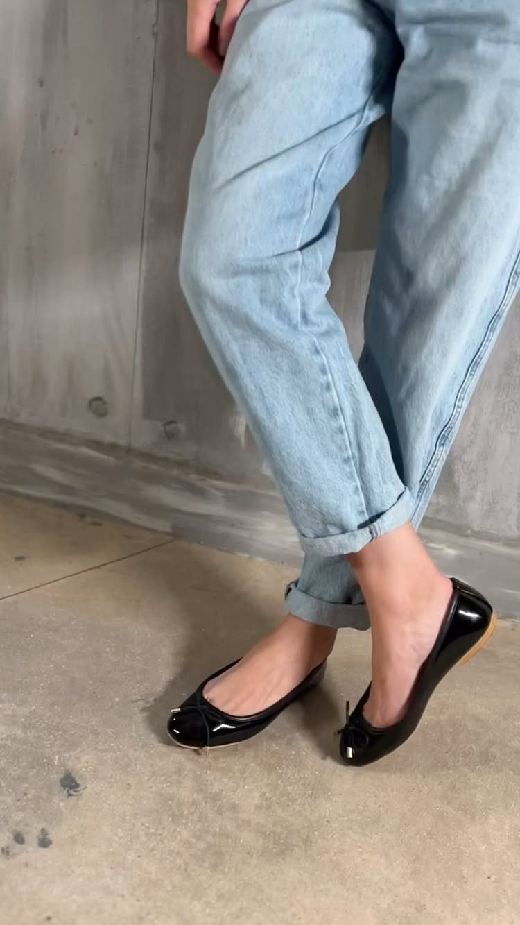 Black patent leather ballet flats worn with light blue denim jeans on a concrete floor.