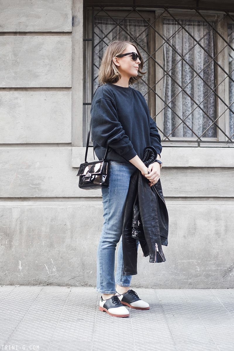 A woman standing on a sidewalk wearing blue jeans and black and white leather oxford shoes.