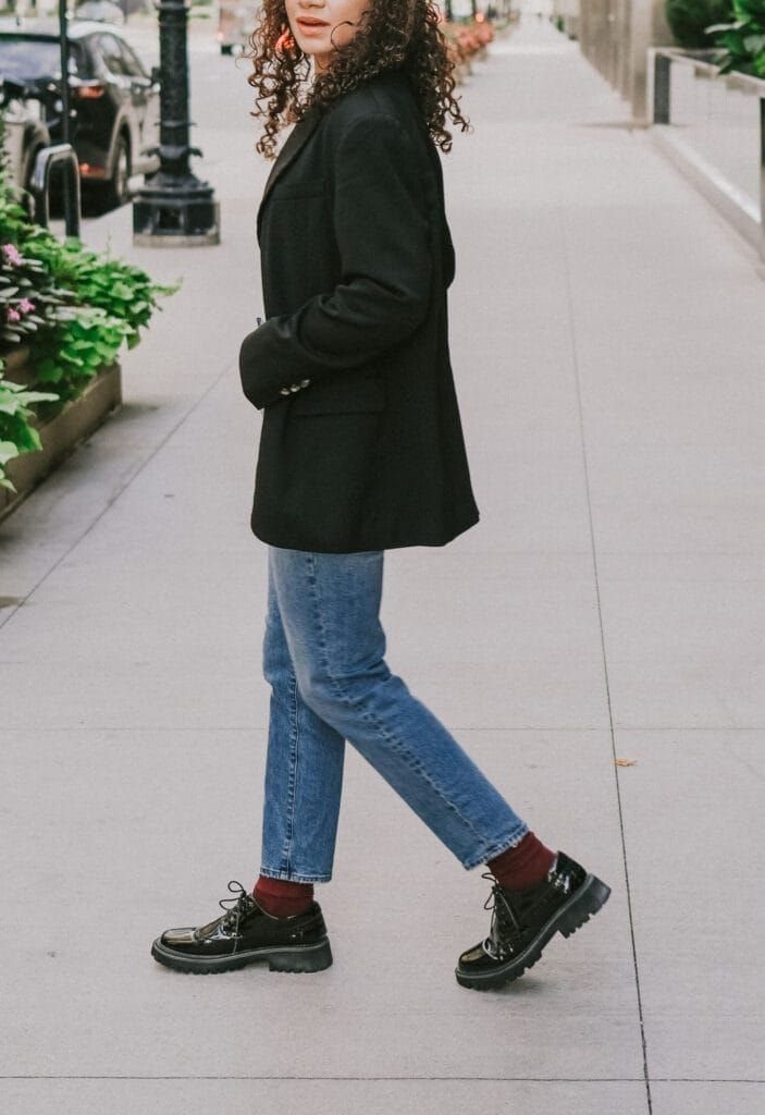 A person walking on a sidewalk wearing blue jeans and chunky black lace-up shoes.