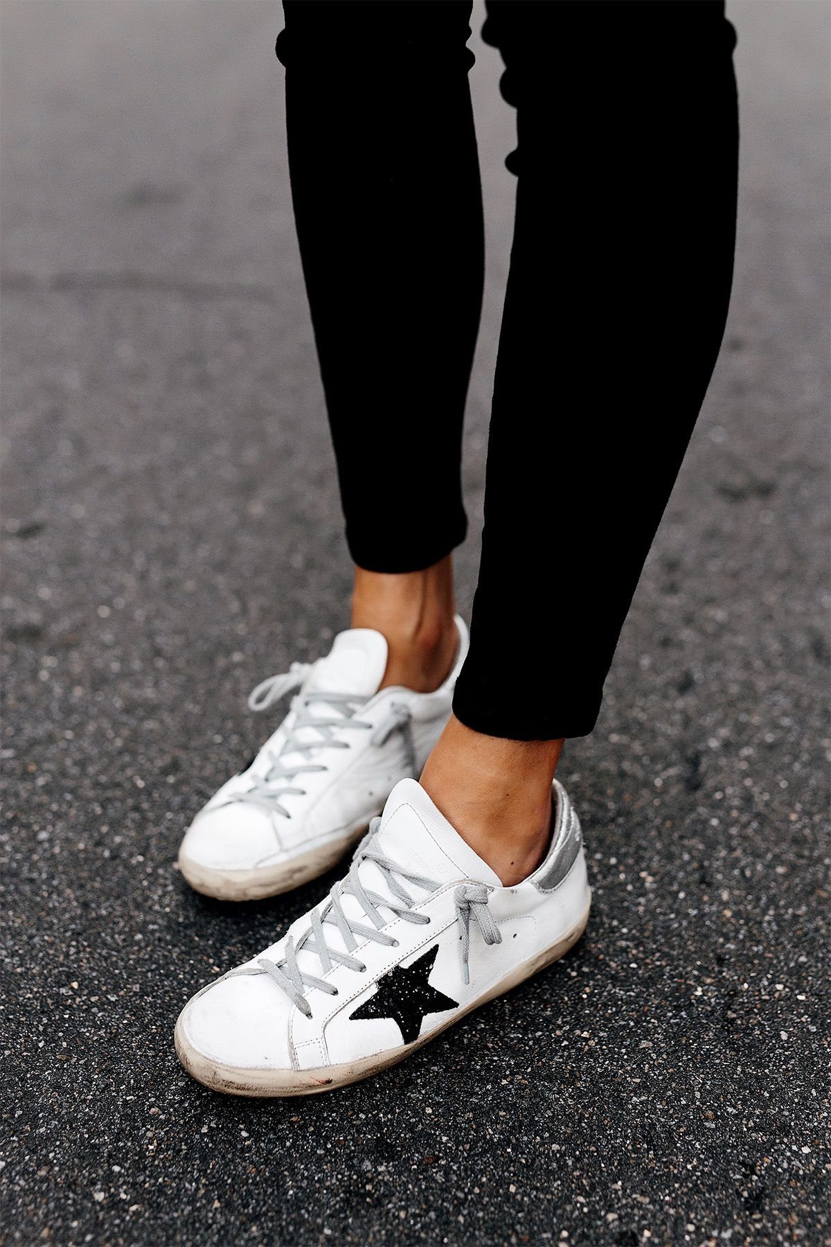 White distressed sneakers with a black star worn with black skinny jeans on asphalt.