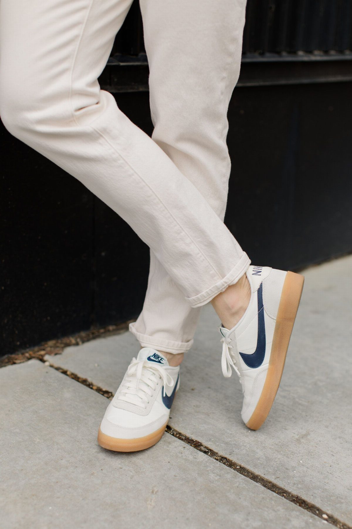 Person wearing cream jeans and white sneakers with navy swoosh and gum soles on a sidewalk.