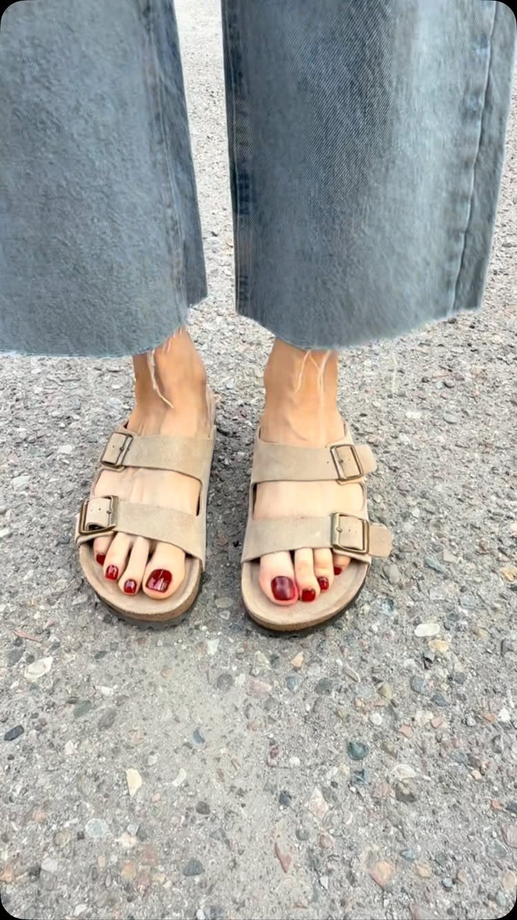 Beige two-strap sandals worn with blue denim jeans on an asphalt ground.