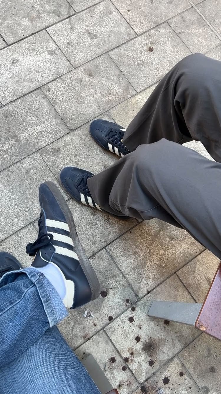 Two people sitting on stone tiles wearing black Adidas sneakers with white stripes and various trousers.