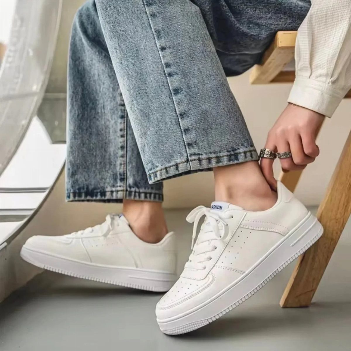 A person wearing blue denim jeans and white low-top sneakers sitting on a wooden stool.