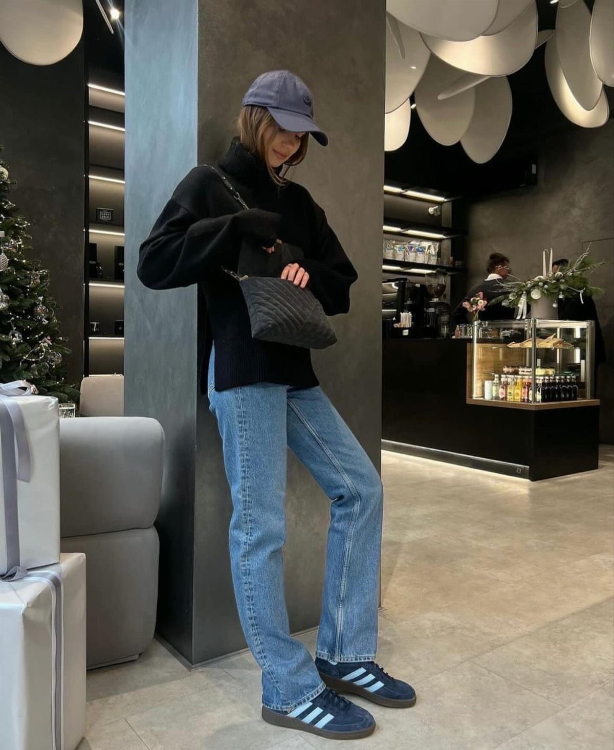 A person standing in a cafe wearing blue jeans and navy blue sneakers with white stripes.