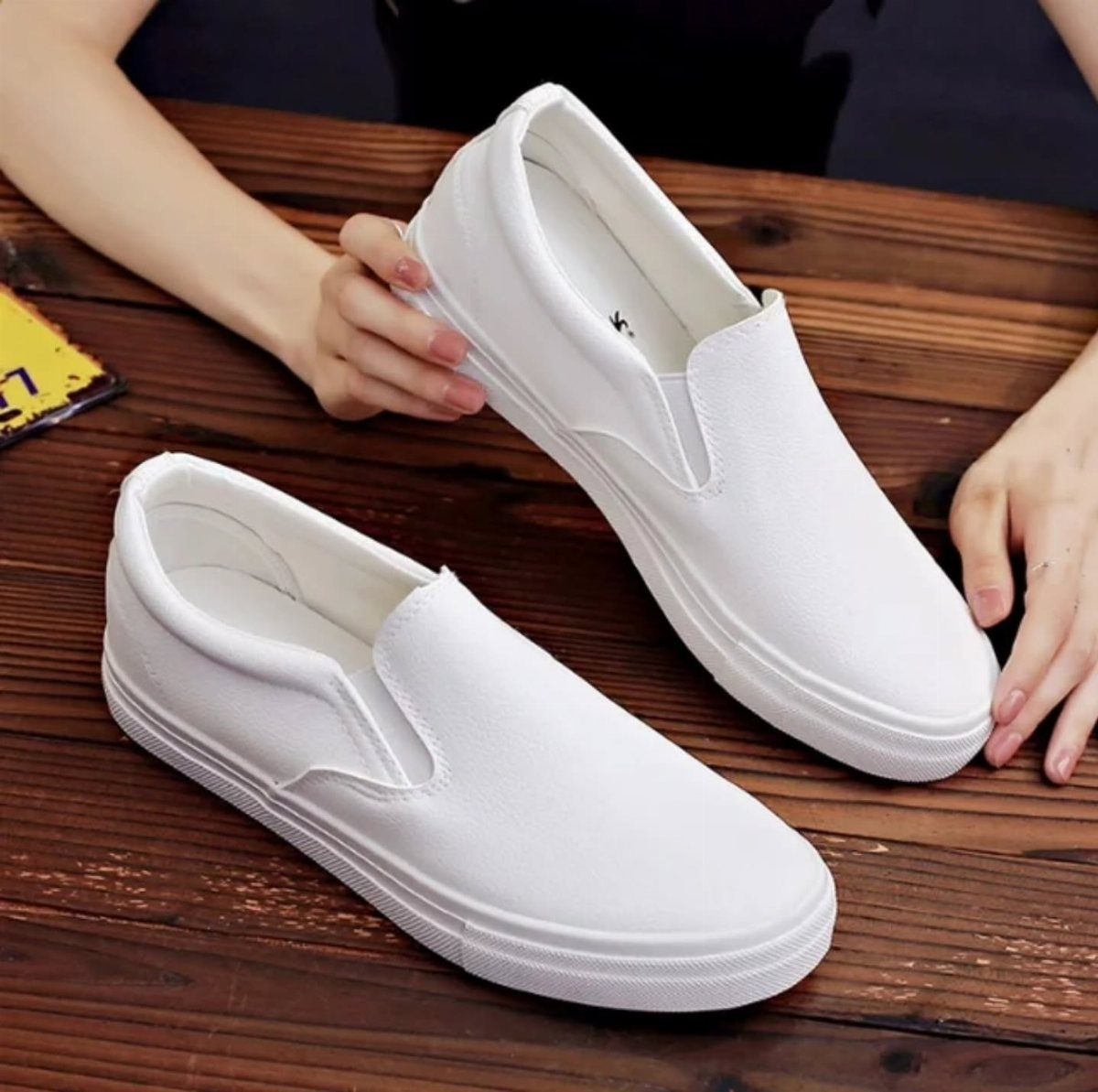 White canvas slip-on shoes being held by hands on a wooden table.