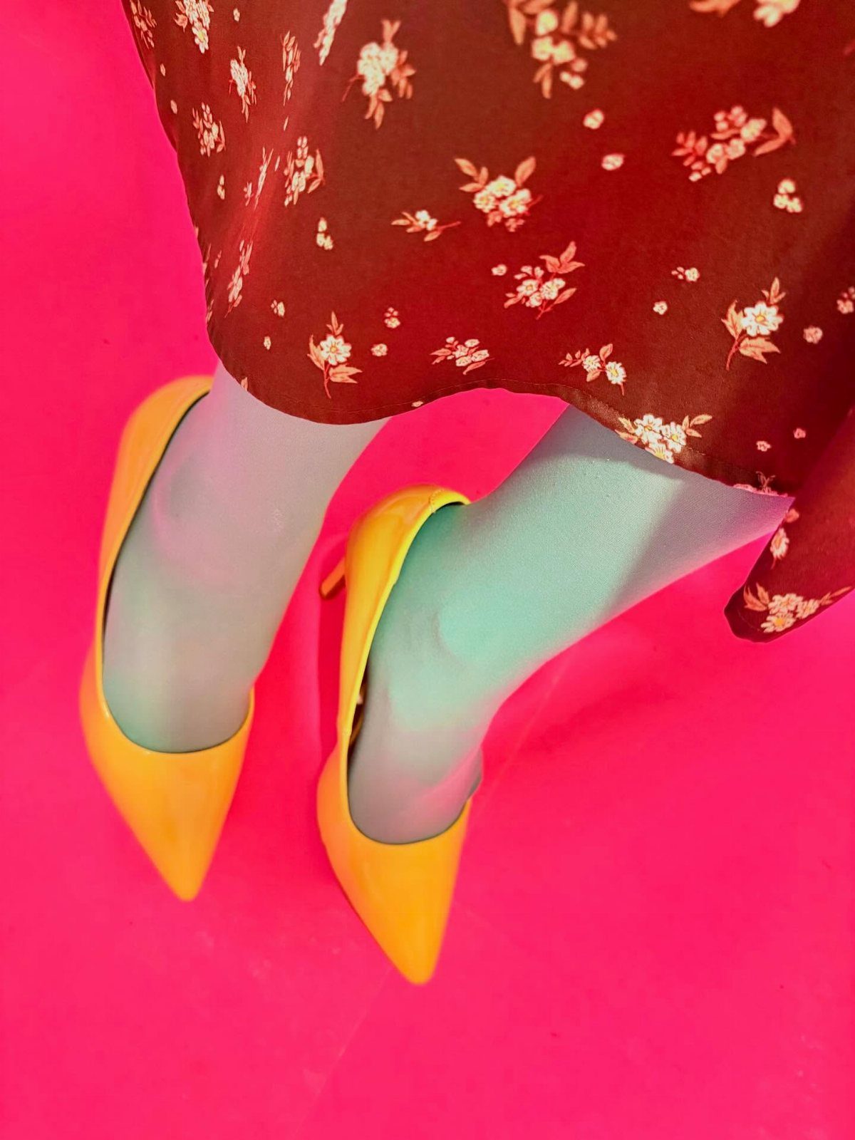 Yellow pointed-toe high heels worn with a brown floral dress against a pink background.