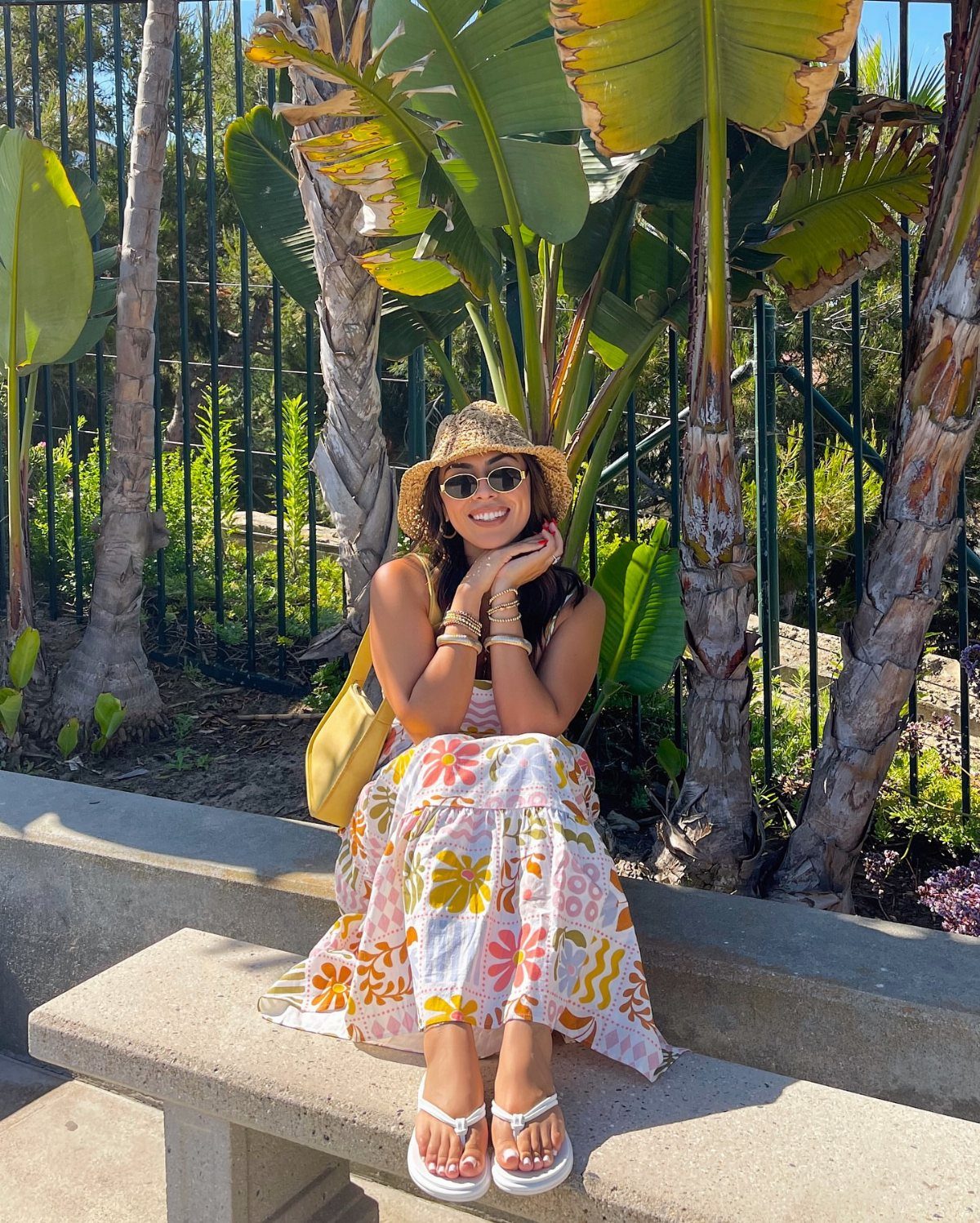 Woman in a floral midi dress and white flip flops sitting on a bench outdoors.