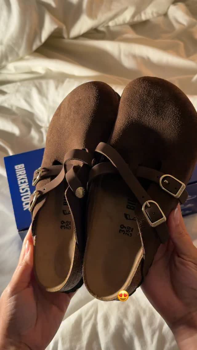 Two hands holding a pair of brown suede Birkenstock clog sandals over white sheets.