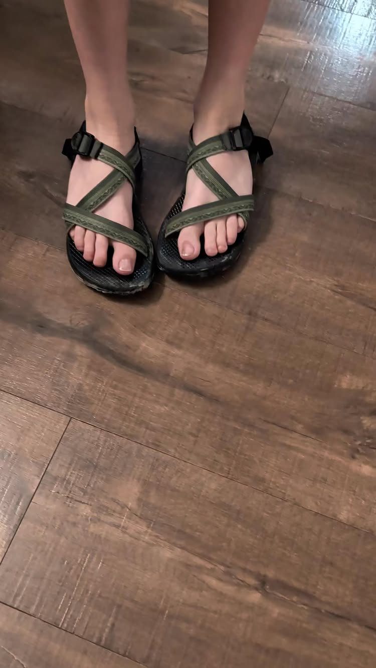 Person wearing dark green sport sandals with thick straps on a wooden floor.