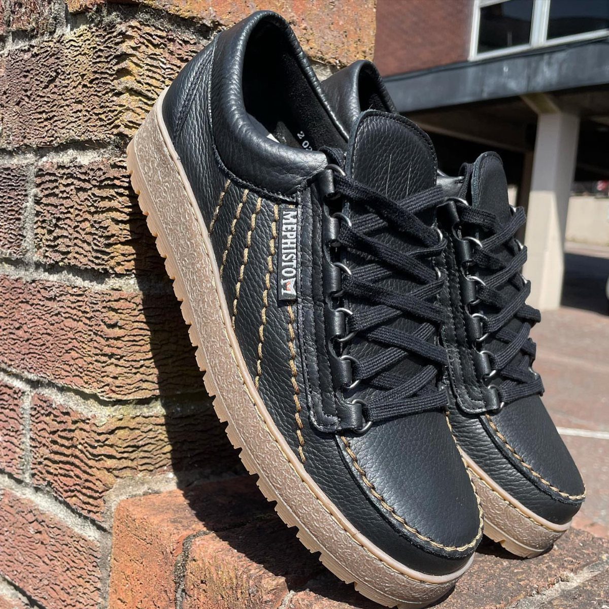 Black leather Mephisto lace-up shoes with tan soles leaning against a brick wall.