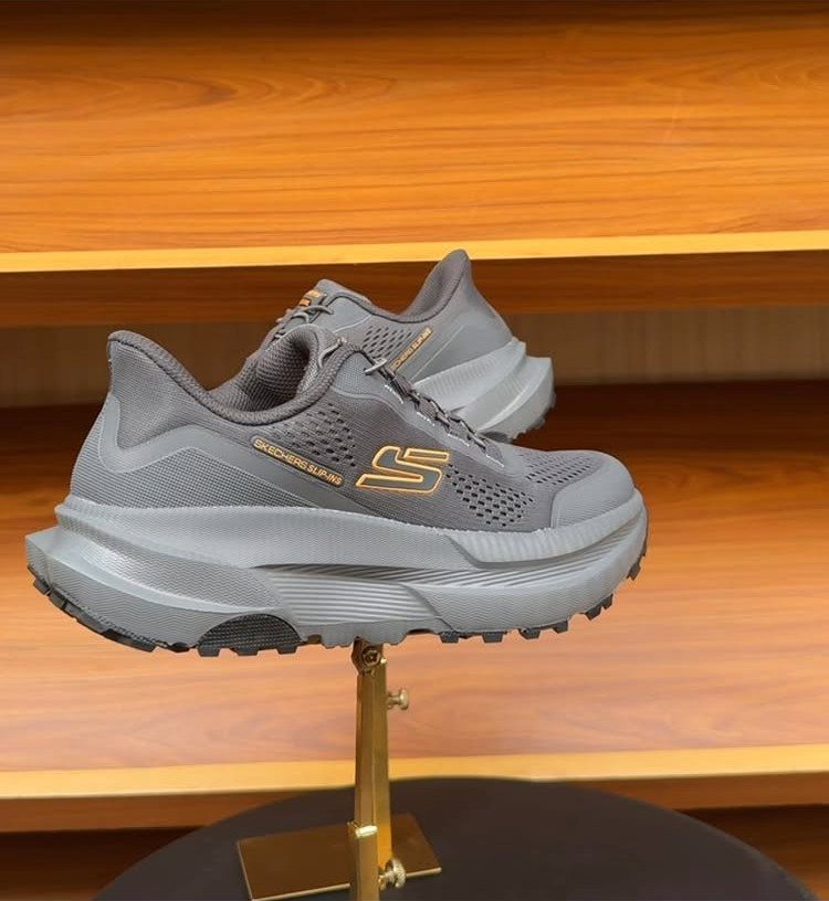 Grey Skechers slip-on athletic shoes resting on a gold display stand in front of wooden shelves.