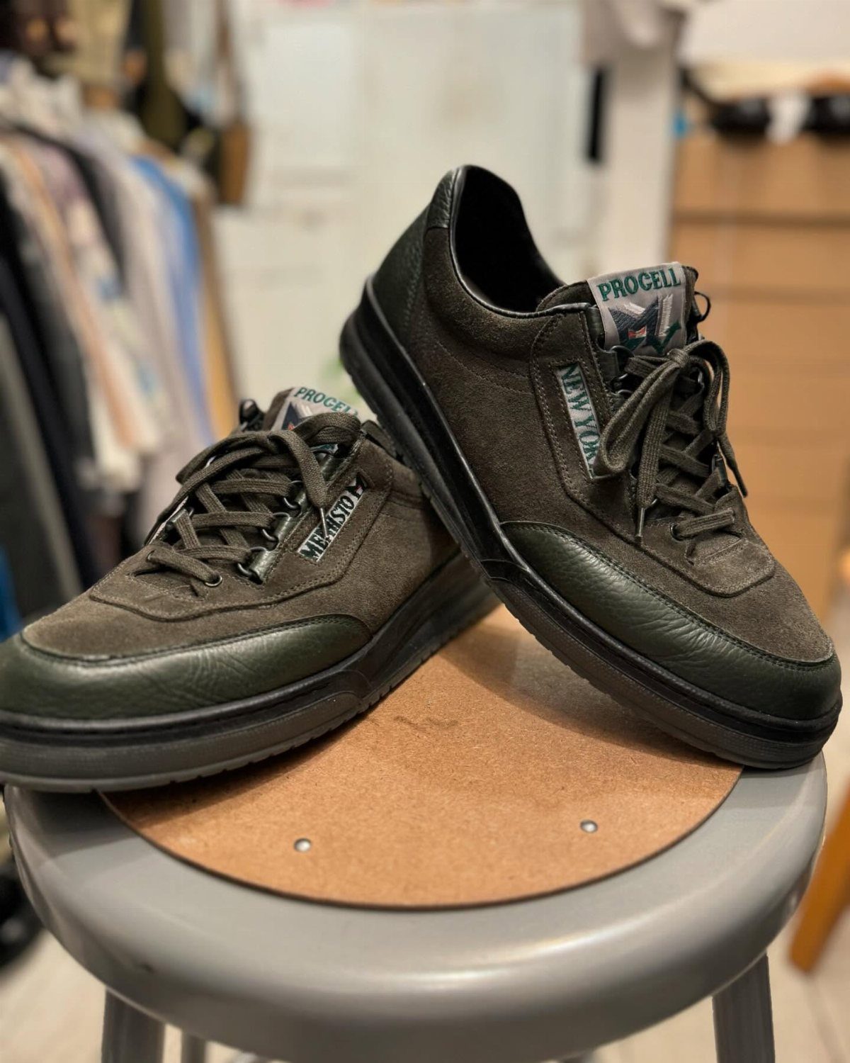 Dark olive green suede sneakers with black laces resting on a small wooden stool.